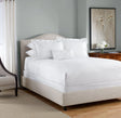 Product display of a hotel queen bed with white luxurious linen dressed on it. Product shows large fluffy pillows and a cloud like duvet. Chair and dress surround the bed.