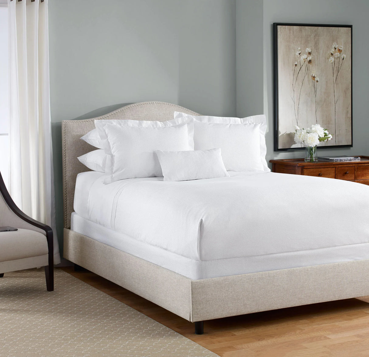 Product display of a hotel queen bed with white luxurious linen dressed on it. Product shows large fluffy pillows and a cloud like duvet. Chair and dress surround the bed.