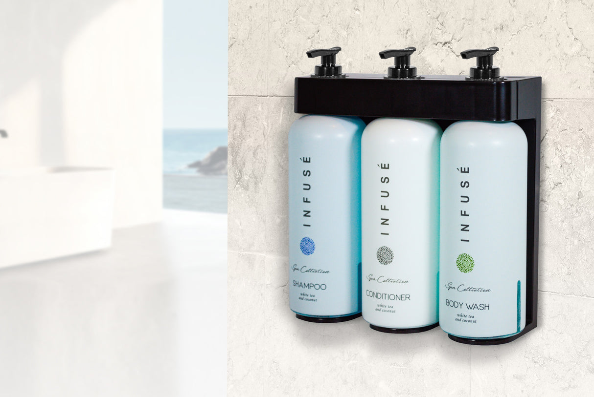 Product shot of Infuse hotel amenities being held by the Aquavera Dispenser holder. Works well with shampoo, conditioner, bodywash and other hotel amenities. Background is a bright bathroom looking over the ocean.
