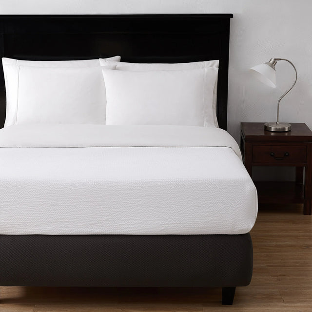 A bed showcasing a while top sheet and pillows. To the right is a nightstand with a lamp on top.