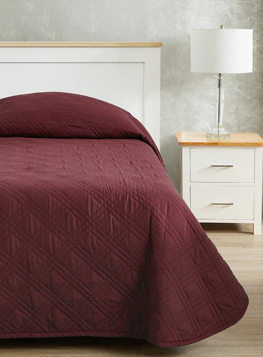 Burgundy red colored bedspread coverlet for single bed. Bed has white frame with wood detailing. Matching nightstand to the right with metal lamp on top.