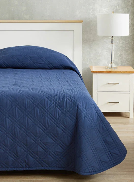 Navy blue colored fitted coverlet for single bed. Bed has white frame with wood detailing. Matching nightstand to the right with metal lamp on top.