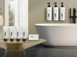 Product shot in bathroom. On right are sample hotel amenities in shampoo, conditioner, body wash, body lotion and cleansing bar. On the right is the larger dispenser version of those products hanging over a modern white bathtub.