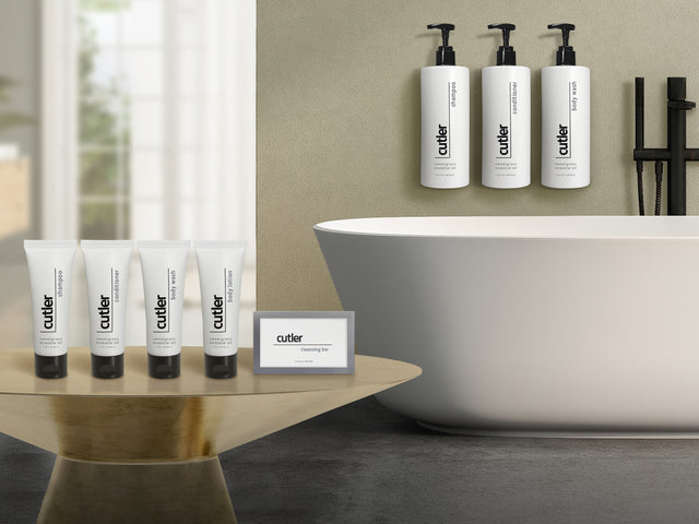 Product shot in bathroom. On right are sample hotel amenities in shampoo, conditioner, body wash, body lotion and cleansing bar. On the right is the larger dispenser version of those products hanging over a modern white bathtub.