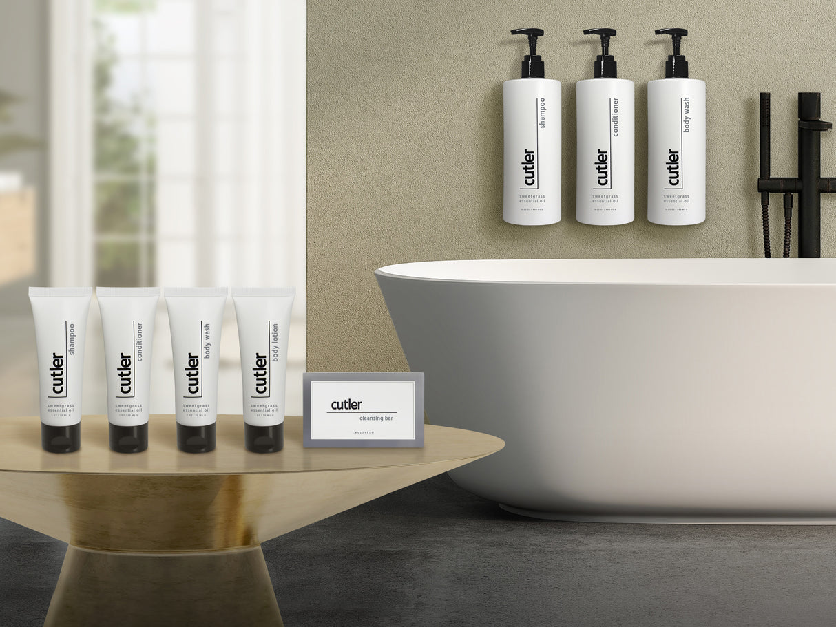Product shot of sample and dispenser hotel amenities. To the left are sample sizes of cutler sweetgrass shampoo, conditioner, body wash and body lotion. They are displayed on a gold table. On the right are dispensers of the same amenities hanging over a white modern bathtub.