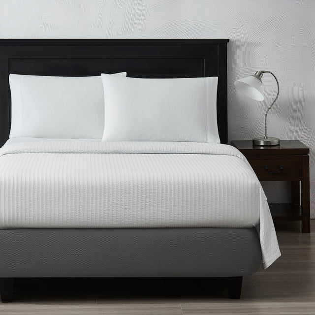 Front of a bed featuring a textured top sheet and fluffy pillows. To the right is a silver table lamp on a night stand.