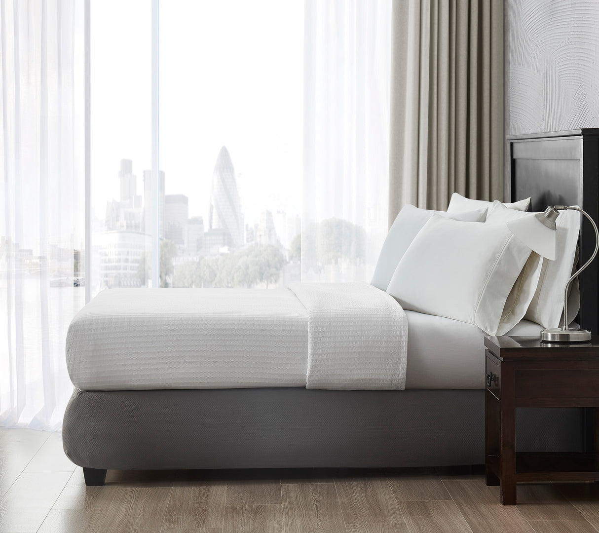 Side view of a bed with a textured top sheet overlooking a metropolitan city. Features a nightstand with a silver lamp on top.