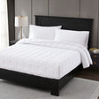 A white down-alternative comforter displayed on a bed with a black headboard, neatly made with pillows and a lamp beside the bed.