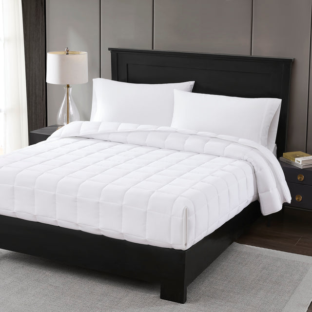 A white down-alternative comforter displayed on a bed with a black headboard, neatly made with pillows and a lamp beside the bed.