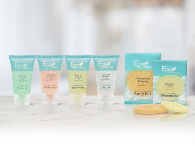 Product layout of the EcoBotanics hotel amenity collection. From left to right we have; White tea and honey products including nourishing shampoo, healthy shine conditioner, refreshing body wash, soothing lotion, moisturizing body bar and cleansing bar. Products are displayed on a marble countertop in a brightly lit room.