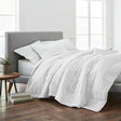 Product display of bed set. Multiple white fluffy pillows and a duvet cover draping over a queen bed. White room with brown floor.