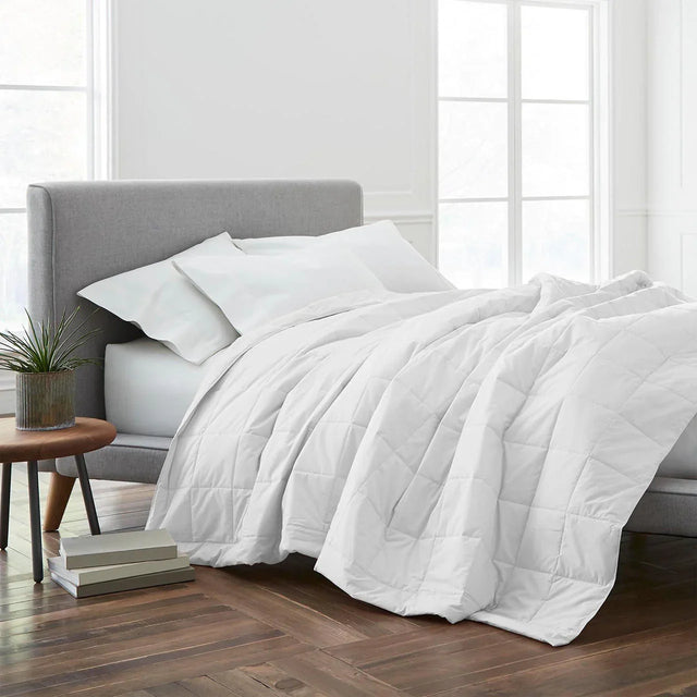Product display of bed set. Multiple white fluffy pillows and a duvet cover draping over a queen bed. White room with brown floor.