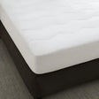A white, quilted mattress pad on a dark-colored mattress, with a zipper or encasement visible on the side.