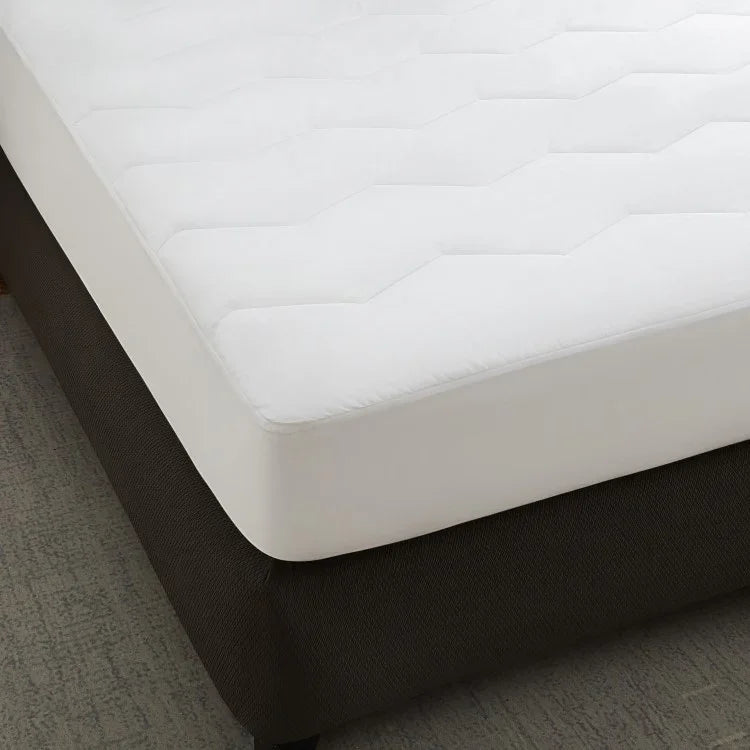A white, quilted mattress pad on a dark-colored mattress, with a zipper or encasement visible on the side.