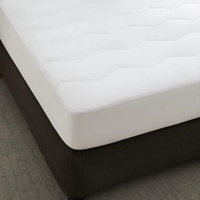 A white, quilted mattress pad on a dark-colored mattress, with a zipper or encasement visible on the side.