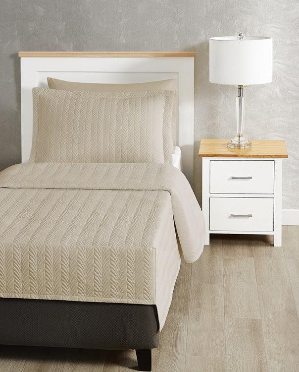 Beige coloured bed sheet coverlet set for a singlebed. White bedframe with wood detailing. Matching nightstand with metal lamp on top.
