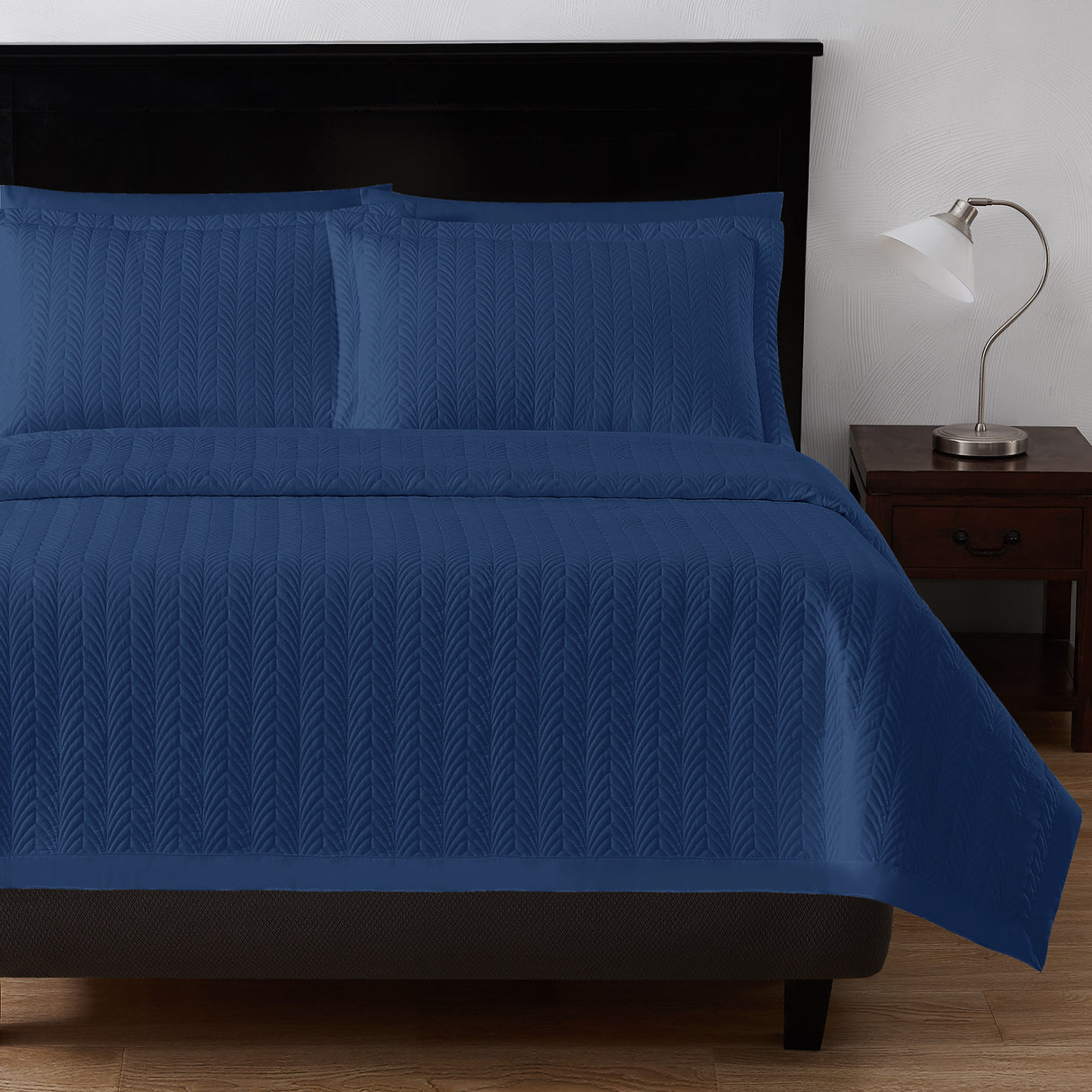 Product shot of leaf textured bed set in  a navy blue color. Image shows the end of the bed along with fluffy pillows. To the right is a nightstand with a lamp on top.