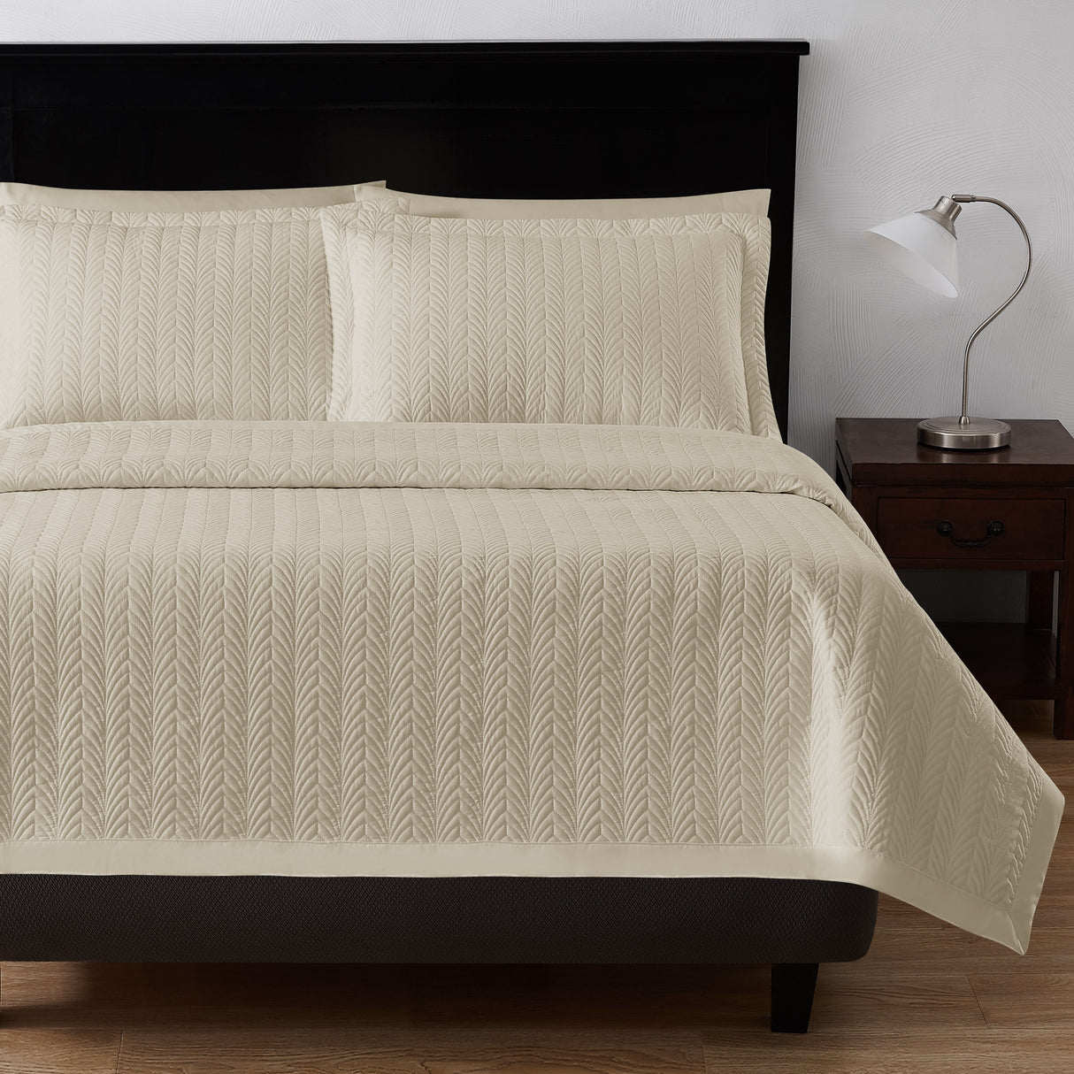 Product shot of leaf textured bed sheet set. Color is a beige/sand color. Image shows end of bed with fluffy pillows. Right is a nightstand with silver lamp on top.