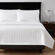 Leaf textured bed set product shot. Image showcases bedset from the end of bed angle along with fluffy pillows. To the right is a nightstand with a lamp on top.