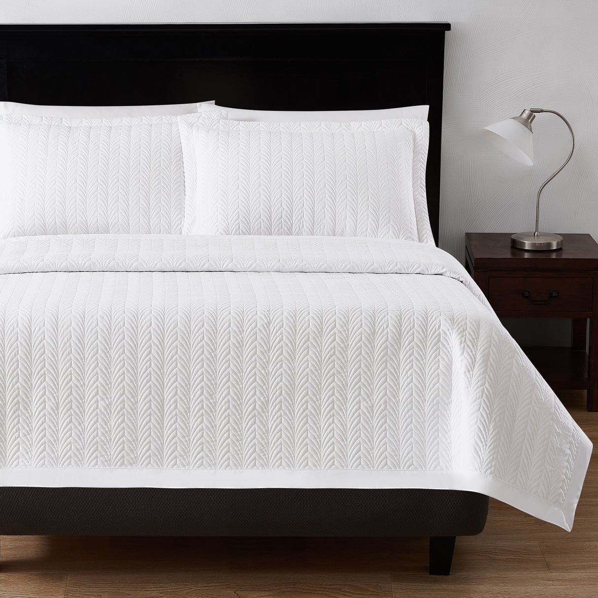 Leaf textured bed set product shot. Image showcases bedset from the end of bed angle along with fluffy pillows. To the right is a nightstand with a lamp on top.