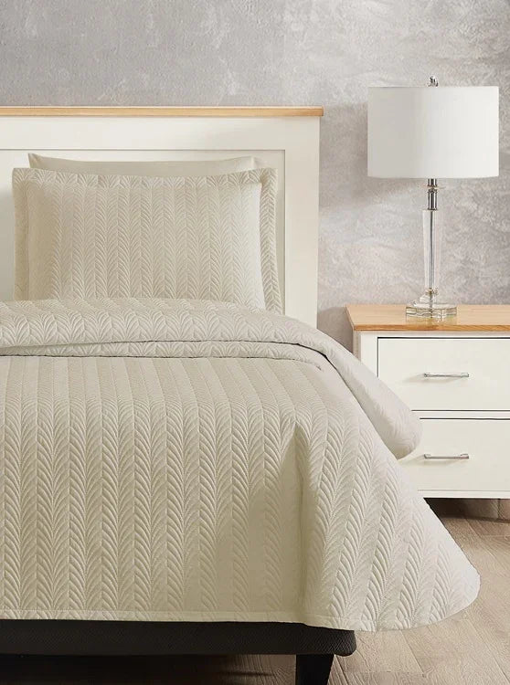 Beige colored bed sheet coverlet set for a single bed. Bottom angle. White bedframe with wood detailing. Matching nightstand with metal lamp on top.