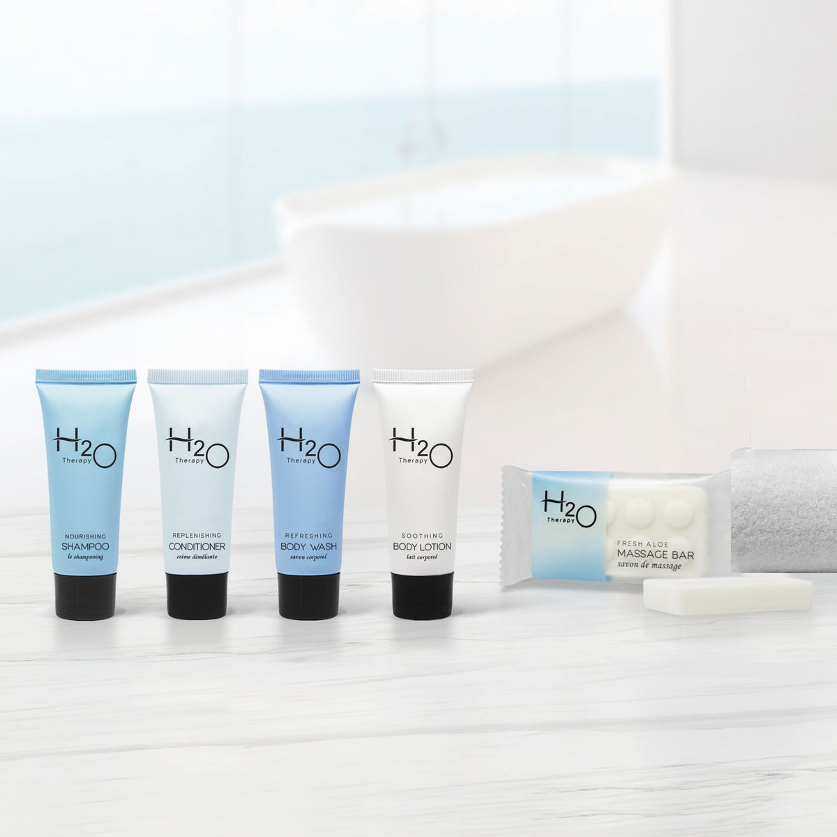 Pictured: H2O Therapy Hotel Amenities line sitting atop a white counter in a white bathroom. The products from left to right are; Nourishing Shampoo, Replenishing Conditioner, Refreshing Body Wash, Soothing Body Lotion, and the Fresh Aloe Massage Bar.