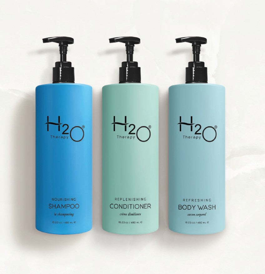 3 bottles of H2O dispenser hotel amenities laying on a marble countertop. From left to right we have; nourishing shampoo, replenishing conditioner and refreshing body wash. Each bottle has a different variety of blue shades to help differentiate.