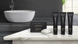 Product display of hotel amenity collection by Infuse. Products showing are lotion, body wash, shampoo and cleansing bar. Products in black packaging with gold detailing. Products are placed on top of marble countertop with a white modern bathtub in the background. Lavender mint scent.