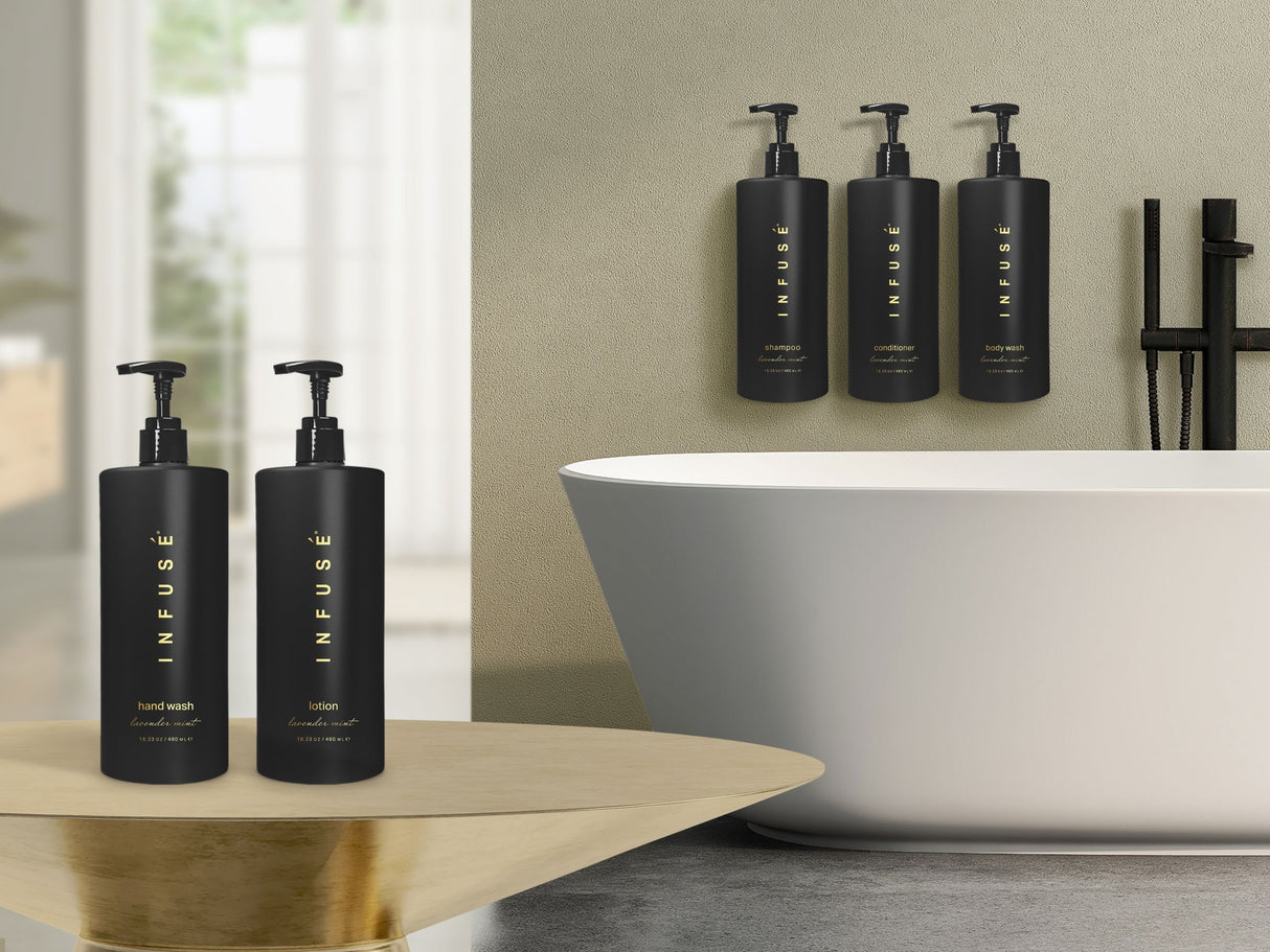 Product shot of Infuse Dispenser hotel amenities. In bathroom products are displayed on gold table including luxurious hand wash, lotion, shampoo, conditioner and body wash. Background is a modern white bathtub.