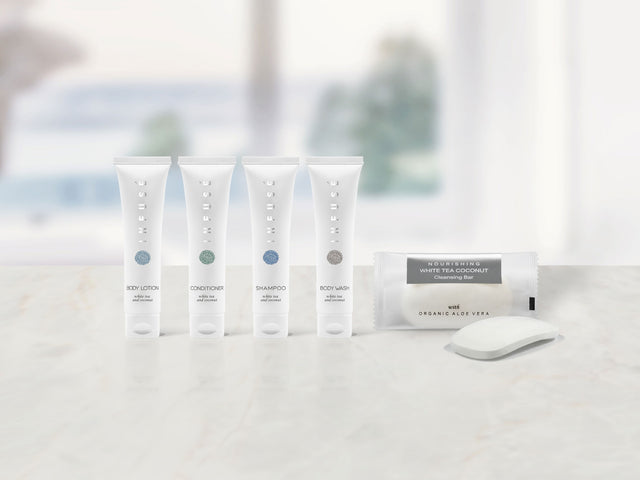 Product shot of the full infuse white hotel amenity collection displayed on a marble countertop. From left to right we have the body lotion, conditioner, shampoo, body wash and cleansing bar.