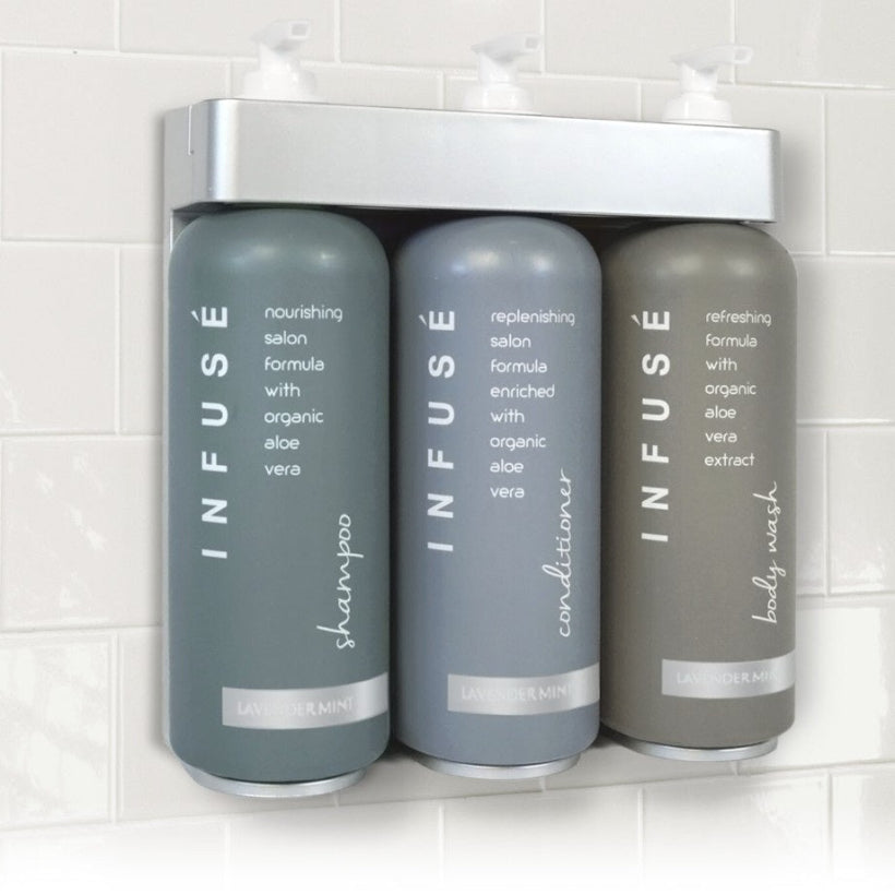 Pictured: Three Infuse collection dispenser bottles hung up in a silver Aquavera Bracket against a white tiled wall. The products from left to right are the shampoo, conditioner, and body wash.