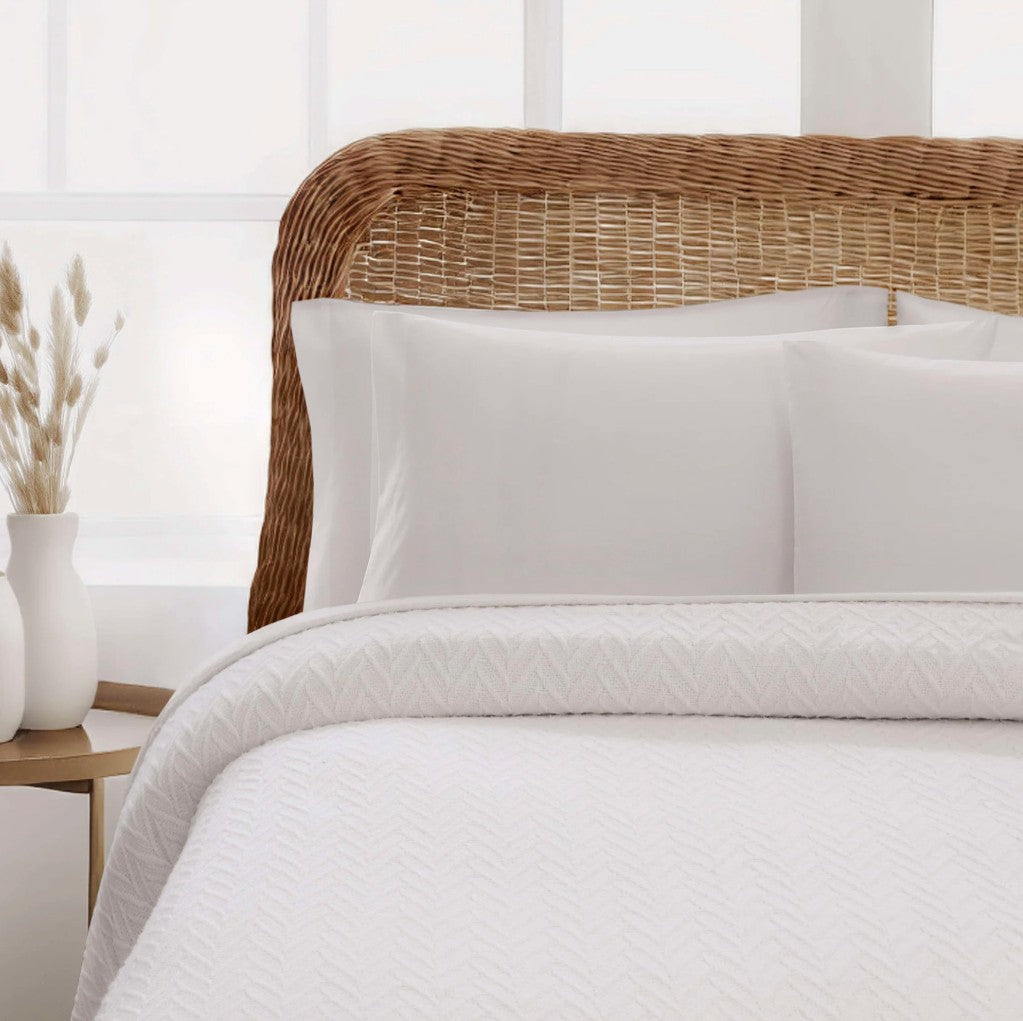 White textured bedding set with two white pillows and a matching white quilted comforter, styled on a bed with a natural wicker headboard in a bright bedroom setting.