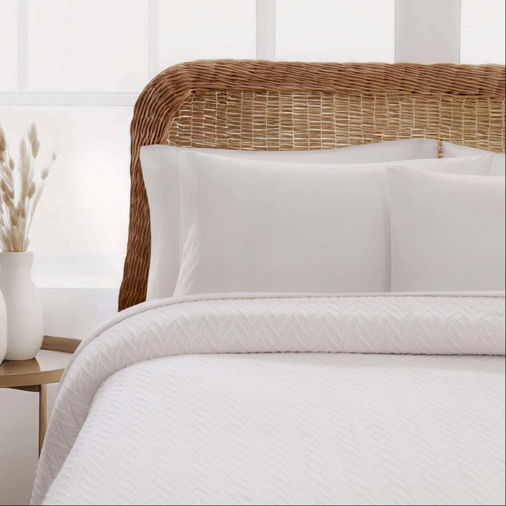 Pictured: White textured bedding set with two white pillows and a matching white quilted comforter, styled on a bed with a natural wicker headboard in a bright bedroom setting.