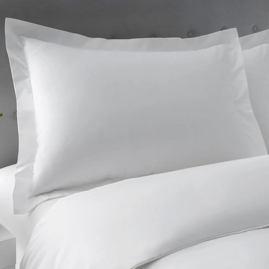 Pictured: Closeup of a pillow on a nicely made white bed against a grey headboard. The pillow is wearing he Millennium T-250 Pillow sham in the Solid White style.
