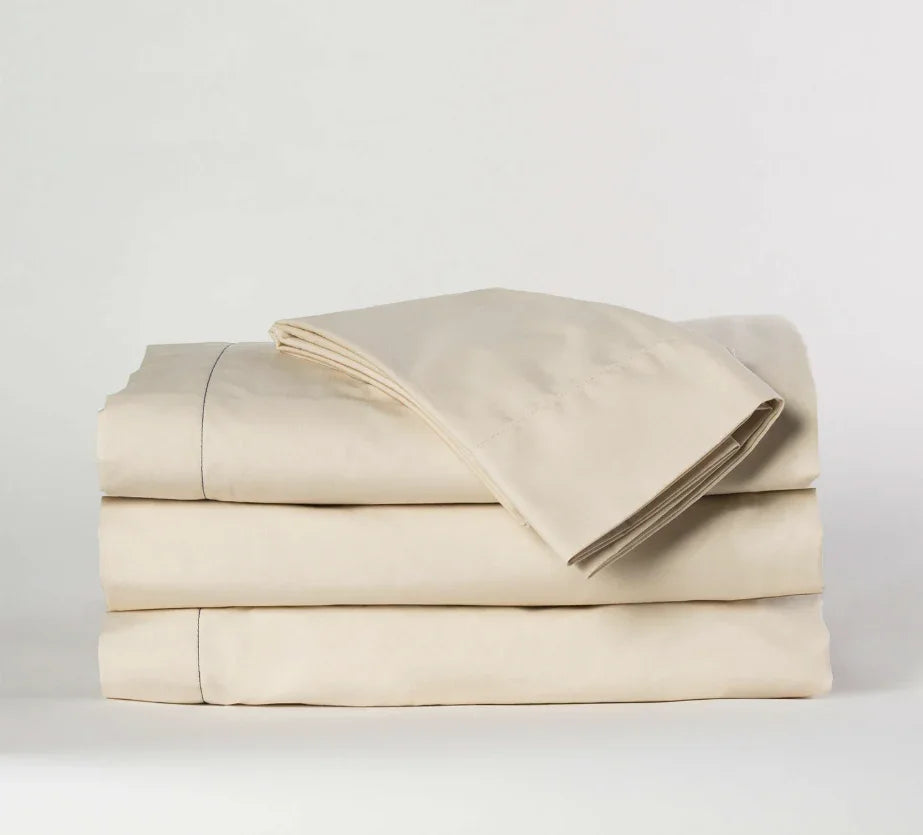 A set of taupe bed sheets on a bed, with a pillow and a nightstand with a lamp in the background.