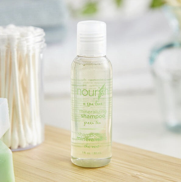 A standing shampoo bottle on a shelf. It says in a green font "Nourish, a spa line, mineralizing shampoo". It is 1 fl. oz. or 30ml. Next to it is a container of q-tips.