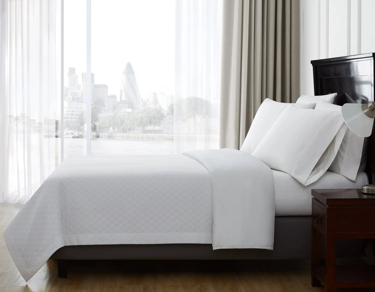 Product shot of a bed cover taken from the side of the bed. Shows fluffy pillows and diamond pattern white bed cover. Background overlooks a metropolitan city.