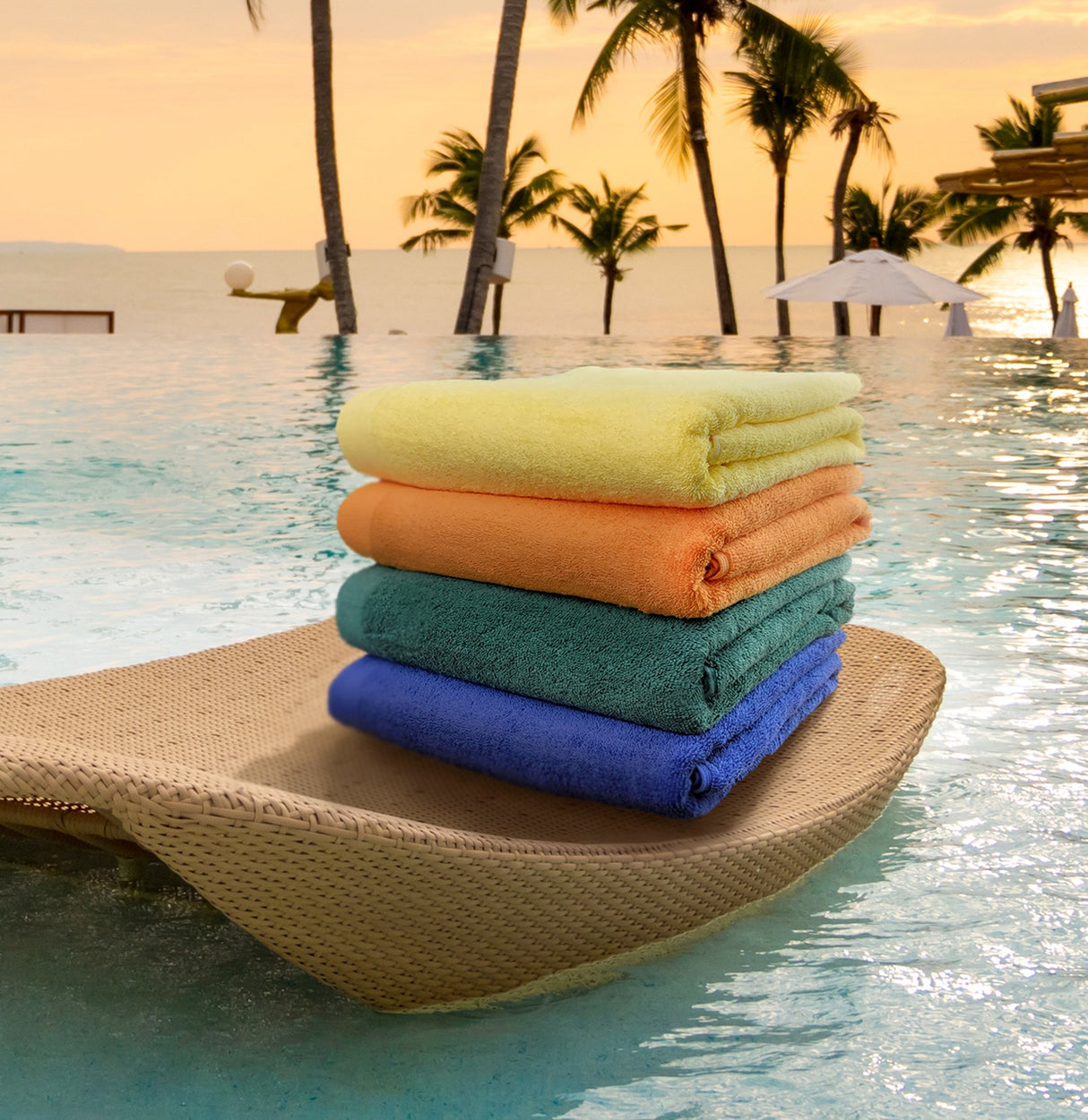 A stack of folded, vibrant colored pool towels placed on a woven mat beside a pool, with palm trees and a sunset in the background.