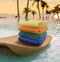 A stack of folded, vibrant colored pool towels placed on a woven mat beside a pool, with palm trees and a sunset in the background.