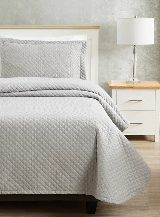 Grey colored bed sheet coverlet set for a single bed. White bedframe with wood detailing. Matching nightstand with metal lamp on top.