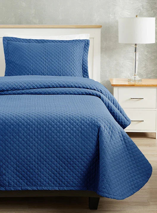 Blue colored bed sheet coverlet set for a single bed. White bedframe with wood detailing. Matching nightstand with metal lamp on top.