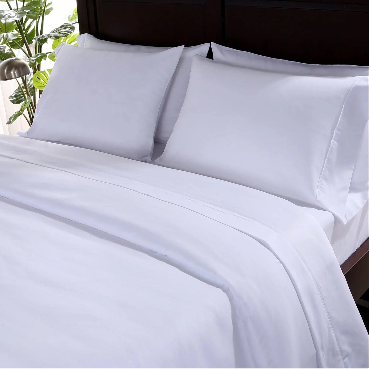 Pictured: Bedroom setting with a white bed on a dark wood frame in the center. This bedding features a top sheet, pillowcases, and a fitted sheet.