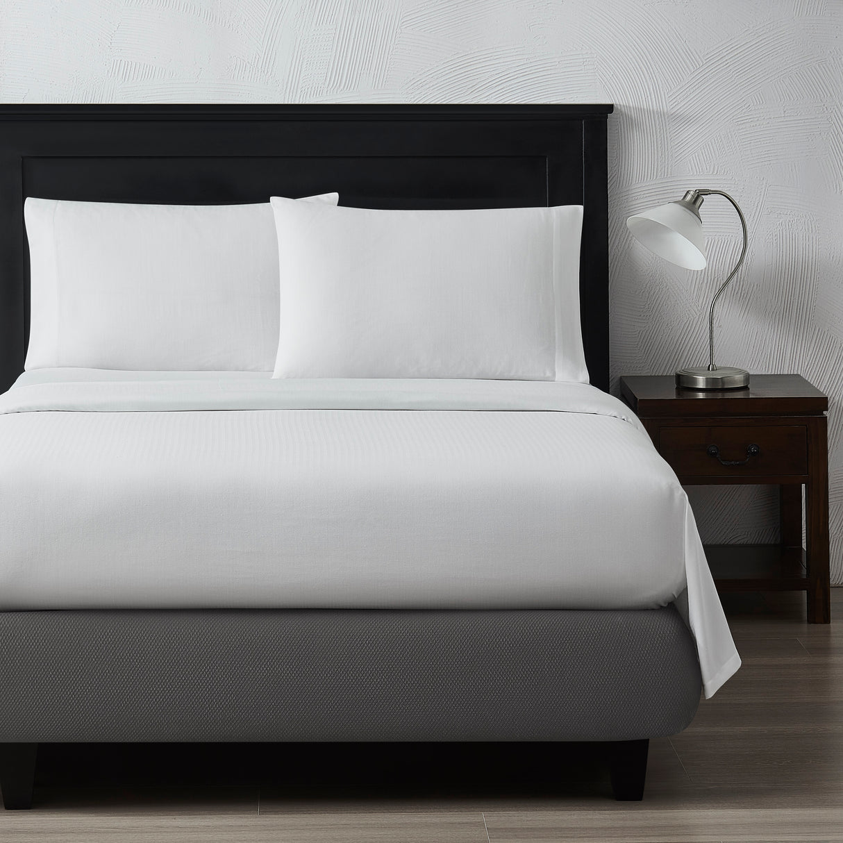 End of a bed showcasing a white top sheet and pillows with a nightstand on the right with a table lamp on top.