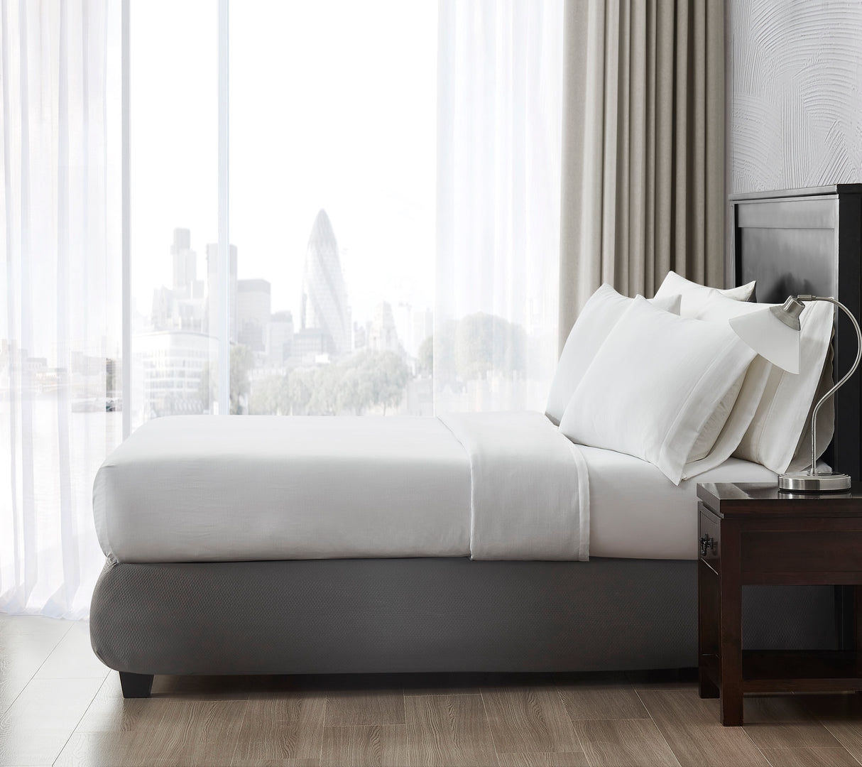 Side view of a bed showcasing a top sheet with pillows near a window overlooking a metropolitan city.