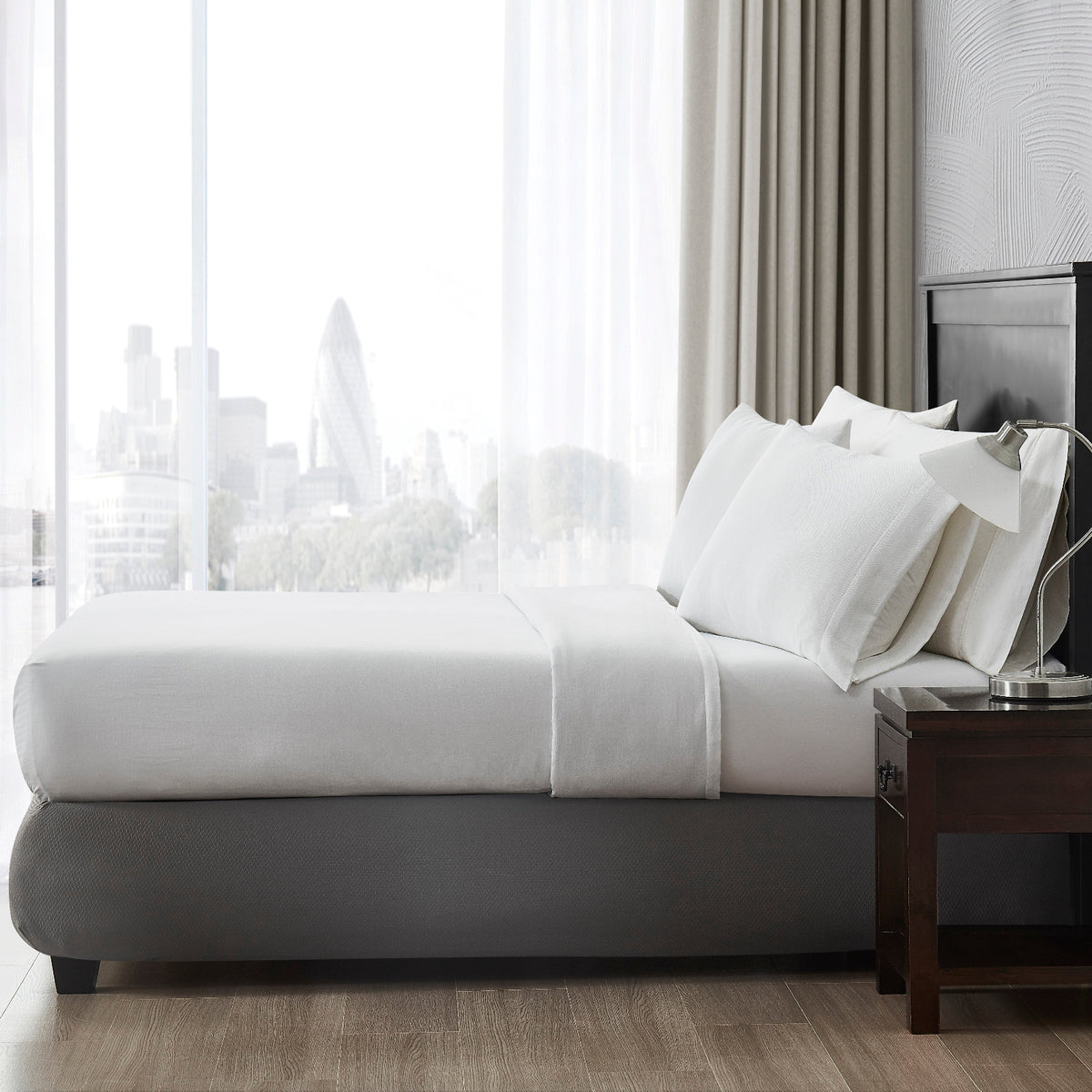 Pictured: Side view of a bed showcasing a top sheet with pillows near a window overlooking a metropolitan city.