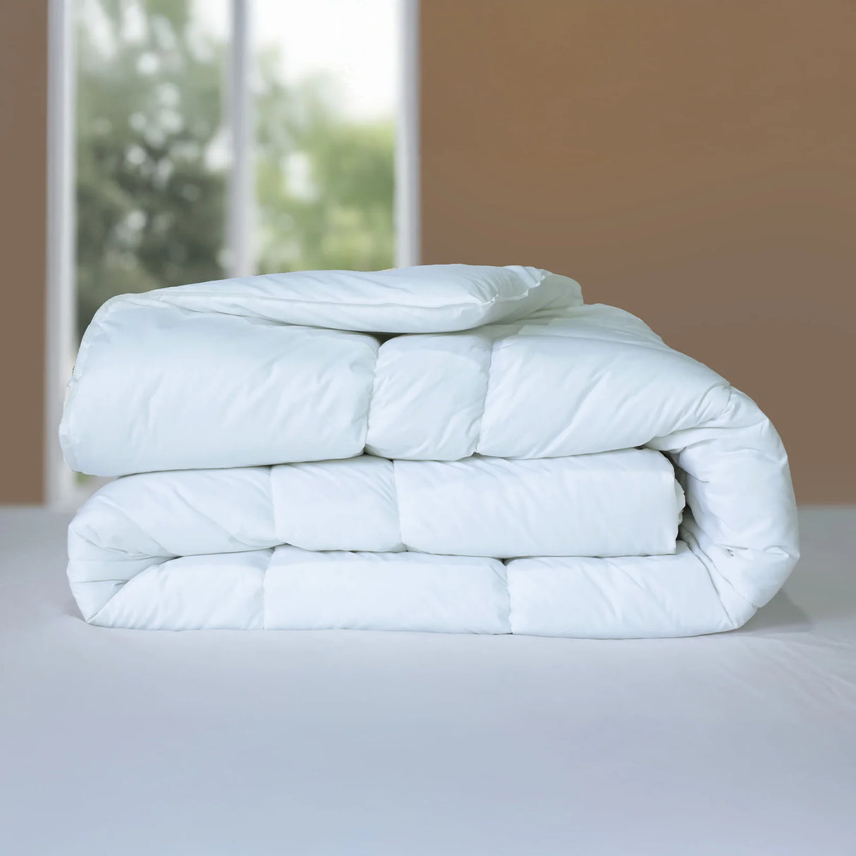 Stack of light blue comforters on a bed with a blurred window background