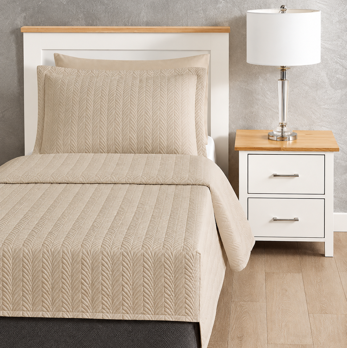 Pictured: Beige bedding on a white wooden bedframe with a matching nightstand to the right holding a glass table lamp with a white shade. The bedding is the Flourish Quilt Coverlet in the shade Sand.