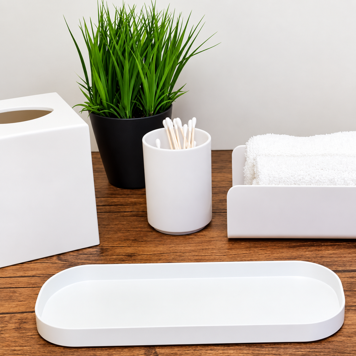 Pictured: A wooden table featuring the Oasis Collection. The Spa Boutique Tissue Box Holder, Apothecary Jar holding cotton swabs, Spa Towel holder with some white towels and the Spa Amenity Tray. There is also a small black cup holding a grass plant.