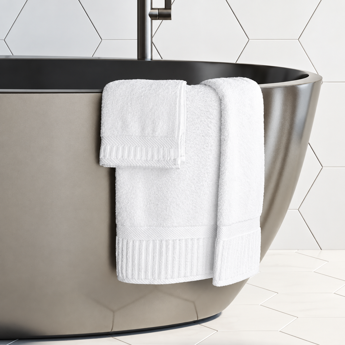 Pictured: Modern white bathroom with honeycomb tile and a stainless steel tub. Draped over the tub are 3 white towels from the Oxford Signature Towel Collection.