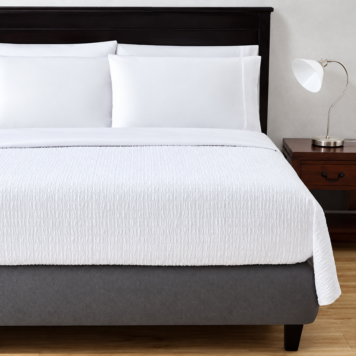 Pictured: Bedroom setting with a dark wood frame and matching nightstand. The white bed features the Jacquard top sheet, which has a naturally scrunched or wrinkled texture in little box stitches.